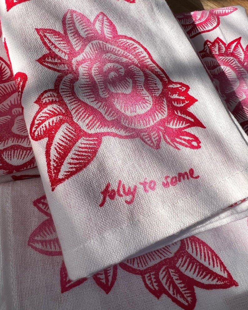 May include: White linen tea towels featuring a repeating pattern of a red rose and leaves. The towels have the handwritten text "jolly to some" in pink. The floral design is a bold, graphic print.