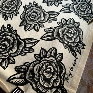 May include: Off-white fabric featuring a repeating pattern of black rose illustrations. Each rose is detailed with intricate lines, and the leaves are also black. The phrase "folly to some" is printed in black script on the fabric.