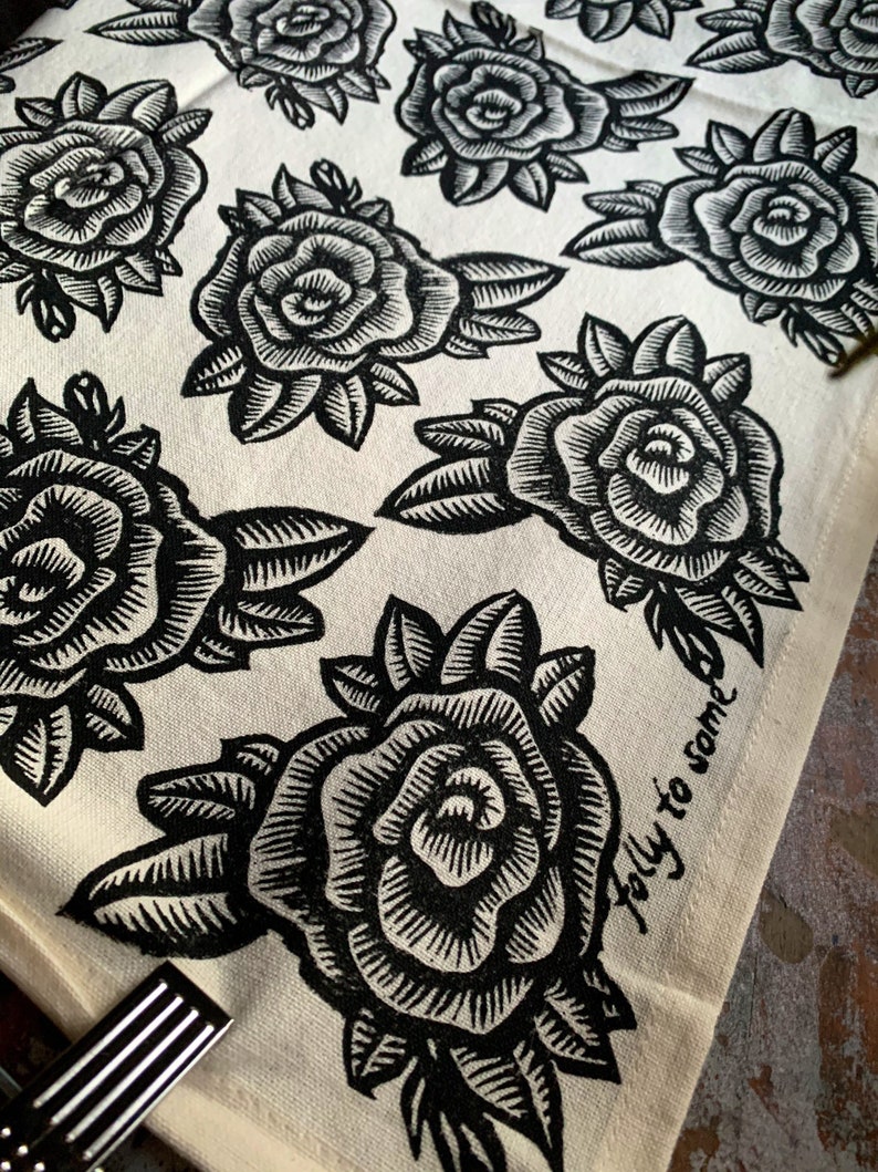 May include: Off-white fabric featuring a repeating pattern of black rose illustrations. Each rose is detailed with intricate lines, and the leaves are also black. The phrase "folly to some" is printed in black script on the fabric.