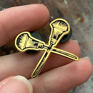 Op de afbeelding: Gouden emaille pin met twee gekruiste kwasten. De pin heeft een zwarte achterkant.