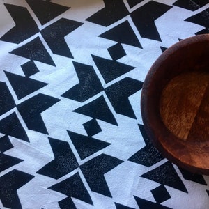 May include: A wooden bowl sits on a white cloth with a repeating black geometric pattern. The bowl is a warm brown color, and the cloth has a textured appearance. The pattern consists of black arrow-like shapes.