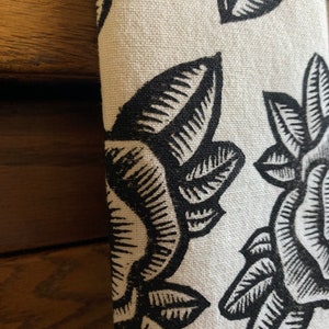 May include: Close-up of folded fabric with a repeating floral pattern. The design features large, stylized black leaves and flowers against a cream background. The fabric has a textured appearance, possibly linen or a similar material.