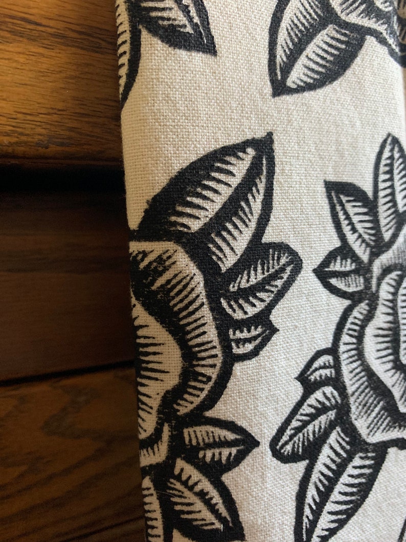 May include: Close-up of folded fabric with a repeating floral pattern. The design features large, stylized black leaves and flowers against a cream background. The fabric has a textured appearance, possibly linen or a similar material.