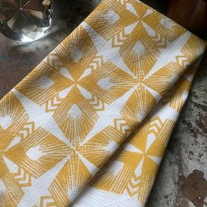 May include: Folded white and gold kitchen towels with a geometric pattern. The towels feature a repeating design of radiating lines and arrow shapes in a mustard yellow color. The towels are folded and stacked on a rustic wooden surface.