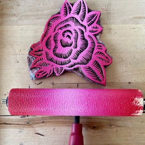 May include: A pink rose-shaped linocut block and a red and pink brayer. The linocut features a detailed floral design with black carved lines. The brayer has a textured surface and a gradient of red to pink.