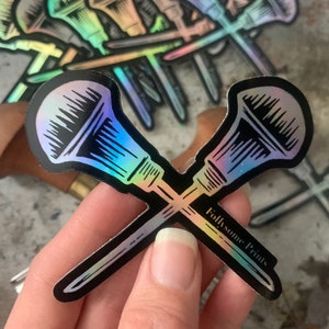 May include: Two iridescent holographic stickers of crossed paintbrushes with black outlines. The stickers are on a wooden surface and are being held by a hand. The text "Follysome Prints" is printed on the bottom of the stickers.
