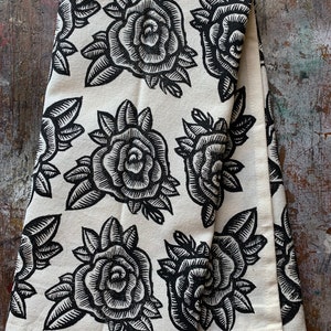 May include: A folded white tea towel featuring a repeating pattern of large, black rose illustrations. The roses are detailed with intricate lines, and the leaves are also black. The tea towel is made of a soft fabric.
