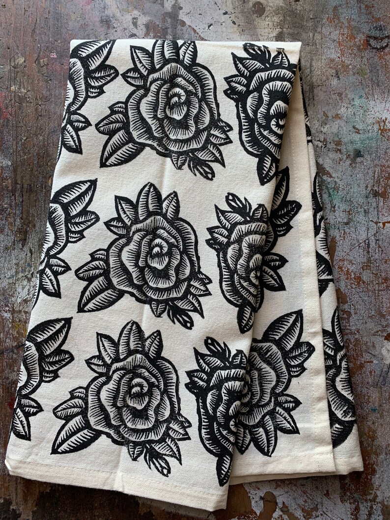 May include: A folded white tea towel featuring a repeating pattern of large, black rose illustrations. The roses are detailed with intricate lines, and the leaves are also black. The tea towel is made of a soft fabric.