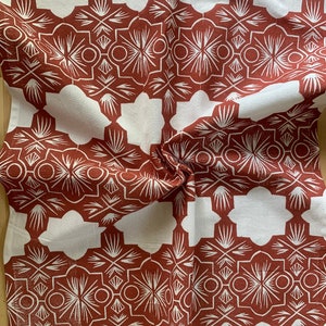 May include: A white and rust-colored patterned textile. The design features a repeating geometric pattern with floral and geometric shapes. The fabric is folded, showing the texture and the interplay of light and shadow across the surface.