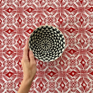 May include: A black and white patterned bowl with a gold rim is held against a red and white patterned background. The bowl's interior features a concentric triangle design. The background has a repeating geometric pattern.