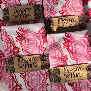 May include: A collection of folded cotton tea towels featuring a repeating rose design in pink and red hues. Each towel is wrapped with a brown paper band, printed with "follysome Prints COTTON TEA TOWEL".