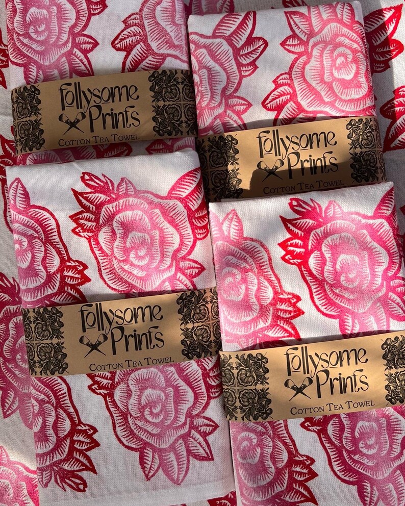 May include: A collection of folded cotton tea towels featuring a repeating rose design in pink and red hues. Each towel is wrapped with a brown paper band, printed with "follysome Prints COTTON TEA TOWEL".