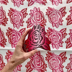 May include: A hand holds a carved wooden rose stamp, with a repeating pattern of red roses on a white fabric background. The stamp is a gradient of pink and red, with detailed lines. The fabric features a similar rose design.