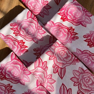 May include: A collection of white napkins featuring a repeating pattern of pink roses and leaves. The roses are printed in a darker pink, contrasting against the white fabric. The napkins are arranged on a wooden surface, likely a tray.