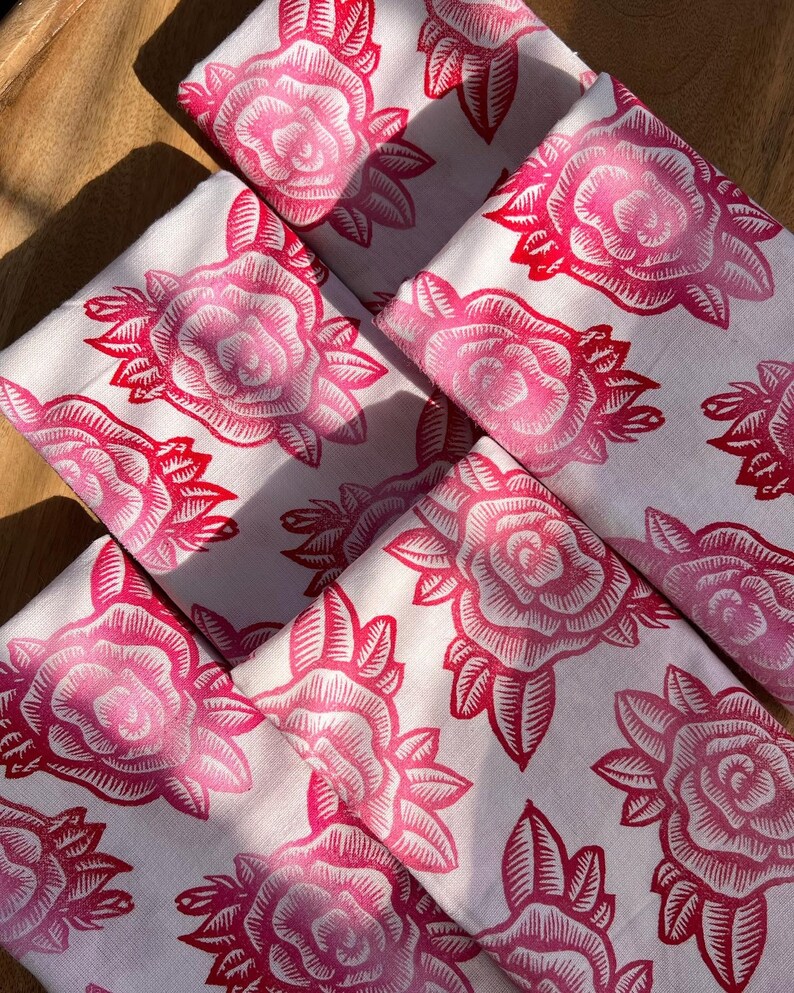 May include: A collection of white napkins featuring a repeating pattern of pink roses and leaves. The roses are printed in a darker pink, contrasting against the white fabric. The napkins are arranged on a wooden surface, likely a tray.