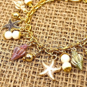 K&ouml;nnte beinhalten: Ein goldenes Kettenarmband mit mehreren Anh&auml;ngern, darunter Muscheln, Seesterne und Perlen. Die Anh&auml;nger sind in verschiedenen Farben, darunter rosa, gr&uuml;n und wei&szlig;.