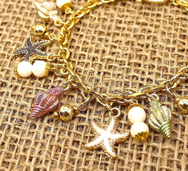 K&ouml;nnte beinhalten: Ein goldenes Kettenarmband mit mehreren Anh&auml;ngern, darunter Muscheln, Seesterne und Perlen. Die Anh&auml;nger sind in verschiedenen Farben, darunter rosa, gr&uuml;n und wei&szlig;.