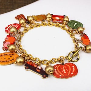 May include: Gold-toned charm bracelet featuring autumn-themed charms. Charms include pumpkins, gourds, and a truck carrying pumpkins. The bracelet has a toggle clasp and gold bead accents.