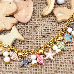 K&ouml;nnte beinhalten: Ein goldenes Kettenarmband mit mehreren Anh&auml;ngern, darunter Muscheln, Seesterne und Perlen. Die Anh&auml;nger sind in verschiedenen Farben, darunter Blau, Gr&uuml;n, Rosa und Wei&szlig;.
