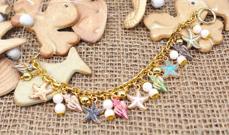 K&ouml;nnte beinhalten: Ein goldenes Kettenarmband mit mehreren Anh&auml;ngern, darunter Muscheln, Seesterne und Perlen. Die Anh&auml;nger sind in verschiedenen Farben, darunter Blau, Gr&uuml;n, Rosa und Wei&szlig;.
