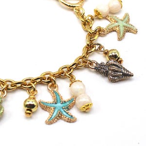 K&ouml;nnte beinhalten: Goldkette Armband mit mehreren Anh&auml;ngern, darunter Muscheln, Seesterne und Perlen. Die Anh&auml;nger sind in Gr&uuml;n-, Blau-, Wei&szlig;- und Goldt&ouml;nen gehalten.