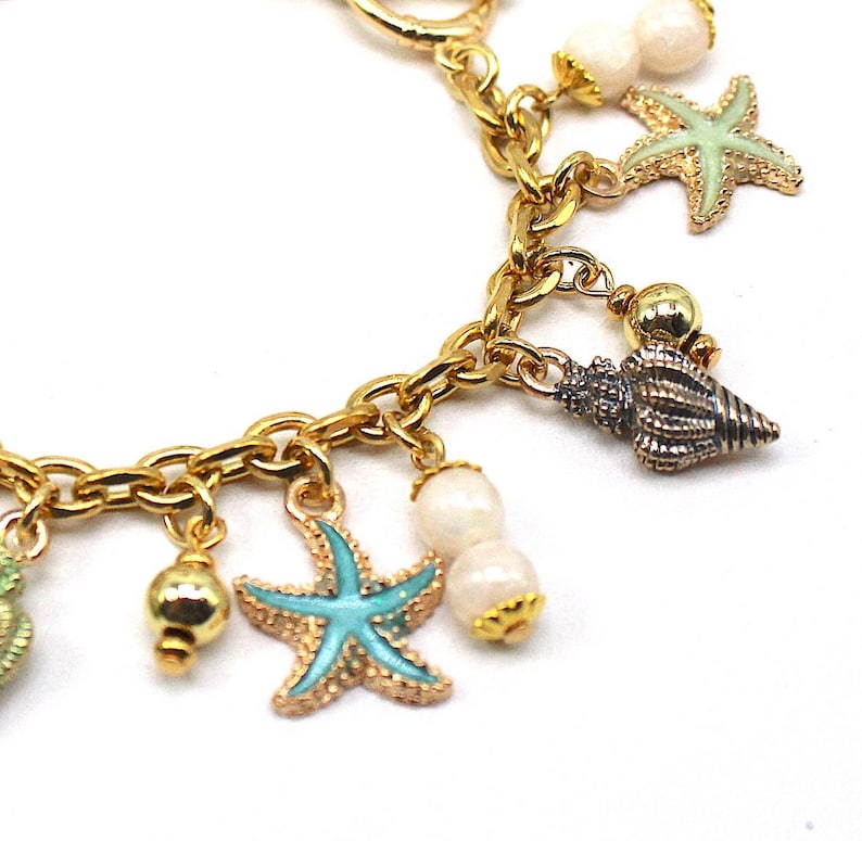 K&ouml;nnte beinhalten: Goldkette Armband mit mehreren Anh&auml;ngern, darunter Muscheln, Seesterne und Perlen. Die Anh&auml;nger sind in Gr&uuml;n-, Blau-, Wei&szlig;- und Goldt&ouml;nen gehalten.