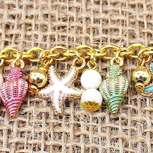 K&ouml;nnte beinhalten: Ein goldenes Kettenarmband mit Anh&auml;ngern in Form von Muscheln und Seesternen. Die Anh&auml;nger sind in verschiedenen Farben bemalt, darunter Pink, Blau, Gr&uuml;n und Wei&szlig;.