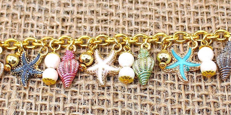 K&ouml;nnte beinhalten: Ein goldenes Kettenarmband mit Anh&auml;ngern in Form von Muscheln und Seesternen. Die Anh&auml;nger sind in verschiedenen Farben bemalt, darunter Pink, Blau, Gr&uuml;n und Wei&szlig;.