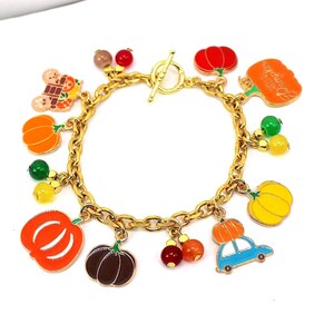 May include: Gold-toned charm bracelet featuring various colorful charms. Charms include pumpkins, a car with a pumpkin on top, and other autumn-themed designs. The bracelet has a toggle clasp.
