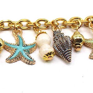 K&ouml;nnte beinhalten: Goldkette Armband mit mehreren Anh&auml;ngern, darunter Muscheln, Seesterne und Perlen. Die Anh&auml;nger sind in Blaut&ouml;nen, Gr&uuml;n und Wei&szlig;.