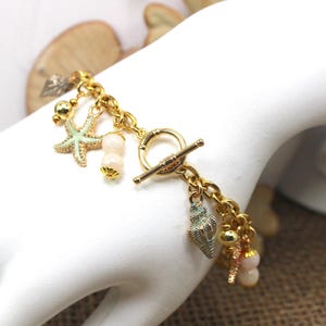 K&ouml;nnte beinhalten: Goldkette Armband mit einem Schiebeverschluss und mehreren Anh&auml;ngern, darunter ein Seestern, Muscheln und Perlen. Die Anh&auml;nger sind in Gr&uuml;n-, Wei&szlig;- und Goldt&ouml;nen gehalten.