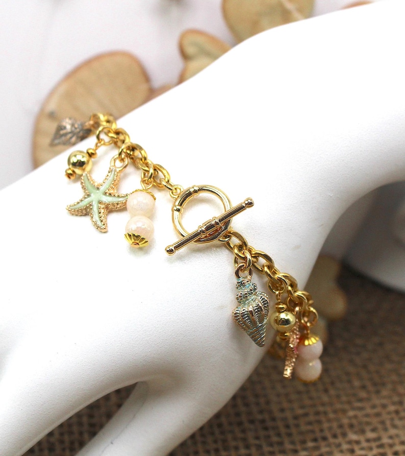 K&ouml;nnte beinhalten: Goldkette Armband mit einem Schiebeverschluss und mehreren Anh&auml;ngern, darunter ein Seestern, Muscheln und Perlen. Die Anh&auml;nger sind in Gr&uuml;n-, Wei&szlig;- und Goldt&ouml;nen gehalten.