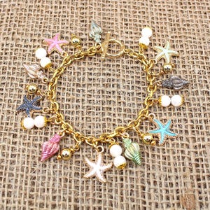 K&ouml;nnte beinhalten: Ein goldenes Kettenarmband mit Anh&auml;ngern in Form von Muscheln, Seesternen und Perlen. Die Anh&auml;nger sind in verschiedenen Farben, darunter rosa, blau, gr&uuml;n und wei&szlig;.