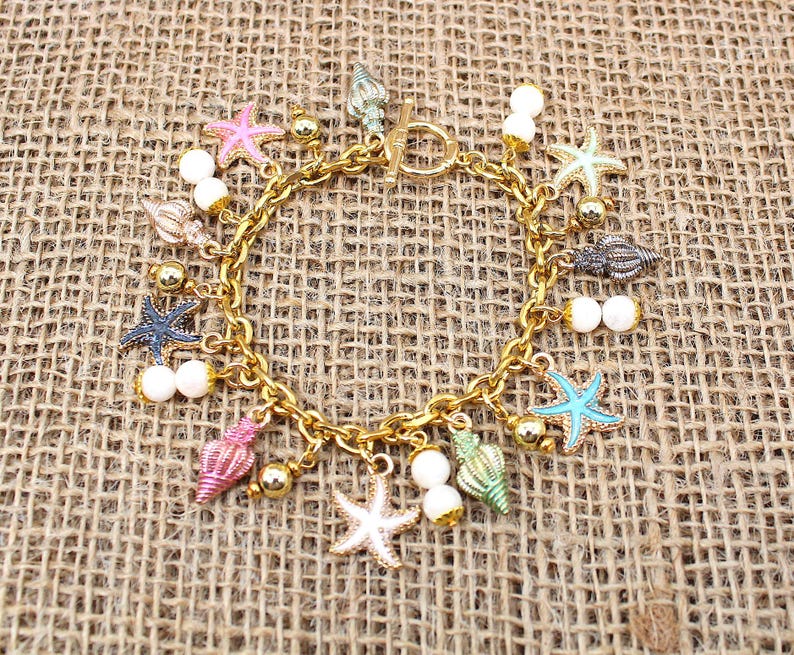 K&ouml;nnte beinhalten: Ein goldenes Kettenarmband mit Anh&auml;ngern in Form von Muscheln, Seesternen und Perlen. Die Anh&auml;nger sind in verschiedenen Farben, darunter rosa, blau, gr&uuml;n und wei&szlig;.