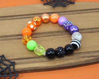 May include: A colorful beaded bracelet with a Halloween theme. The bracelet features a variety of beads in orange, purple, green, black, and clear. Some beads have white polka dots. The bracelet is on a wooden surface with a spider web decoration.