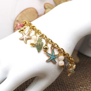 K&ouml;nnte beinhalten: Ein goldenes Kettenarmband mit Anh&auml;ngern in Form von Muscheln und Seesternen. Die Anh&auml;nger sind in Blaut&ouml;nen, Gr&uuml;n- und Wei&szlig;t&ouml;nen bemalt.