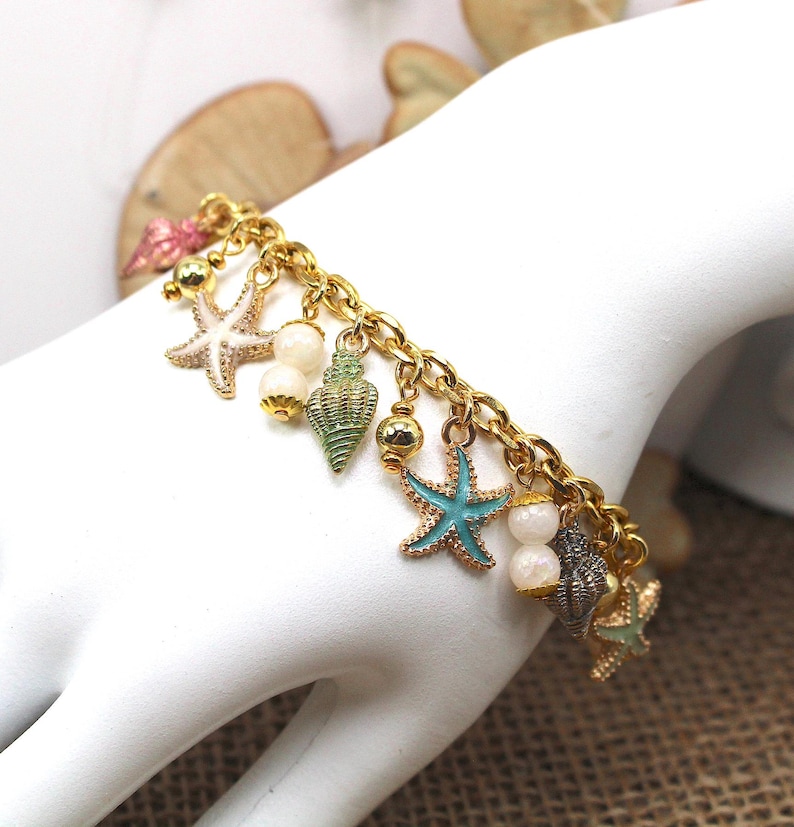 K&ouml;nnte beinhalten: Ein goldenes Kettenarmband mit Anh&auml;ngern in Form von Muscheln und Seesternen. Die Anh&auml;nger sind in Blaut&ouml;nen, Gr&uuml;n- und Wei&szlig;t&ouml;nen bemalt.