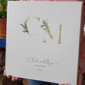 Peut inclure: Livre d'or de mariage blanc avec des initiales "CM" en paillettes dorées entourées de feuillage vert et le texte "Charlie et Meg 23 juillet 2021".