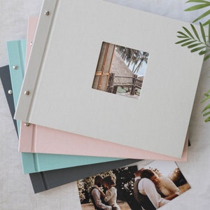 May include: A stack of four photo albums in different colors, including gray, pink, blue, and dark gray. The top album has a window cutout with a photo of a tropical scene. The albums are made of a textured fabric material.