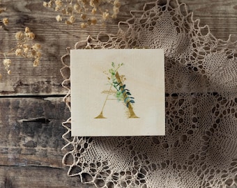 May include: A square, off-white canvas features a gold-colored letter "A" adorned with green and blue botanical accents. The canvas rests on a wooden surface, with a beige lace doily and dried flowers in the background.