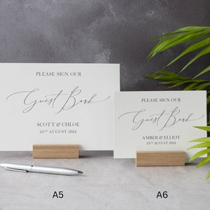 May include: Two white cards with black text on wooden stands. The top card says "Please sign our Guest Book" with the names "Scott & Chloe" and the date "24th August 2024". The bottom card says "Please sign our Guest Book" with the names "Amber & Elliot" and the date "24th August 2024".