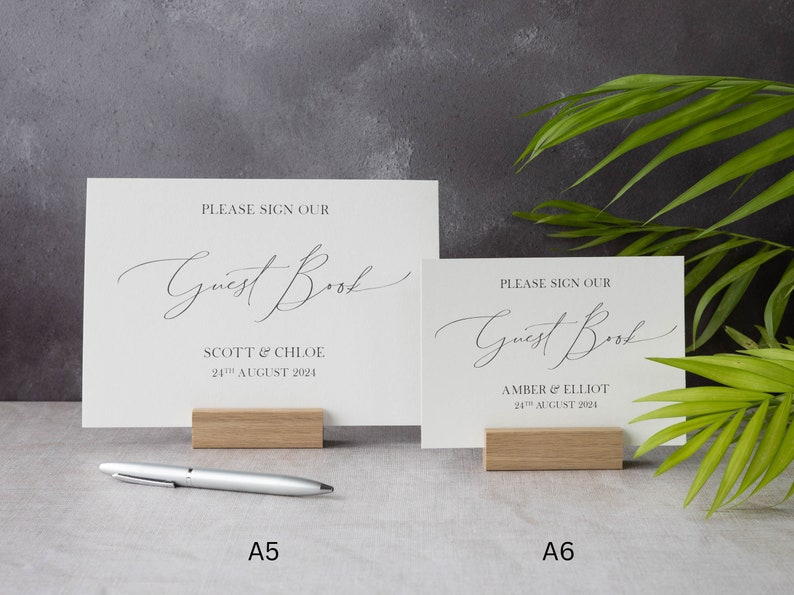 May include: Two white cards with black text on wooden stands. The top card says "Please sign our Guest Book" with the names "Scott & Chloe" and the date "24th August 2024". The bottom card says "Please sign our Guest Book" with the names "Amber & Elliot" and the date "24th August 2024".