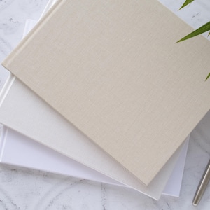 Op de afbeelding: Drie notitieboekjes gestapeld op elkaar. Het bovenste notitieboekje is beige met een linnen textuur. Het middelste notitieboekje is wit met een gladde textuur. Het onderste notitieboekje is wit met een gladde textuur. Een zilveren pen ligt op de tafel naast de notitieboekjes.