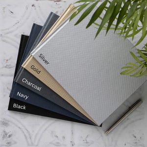 May include: A stack of five notebooks in different colours, including black, navy, charcoal, gold, and silver. The notebooks are arranged in a fan shape, with the silver notebook on top. The notebooks have a textured fabric cover and a simple design.
