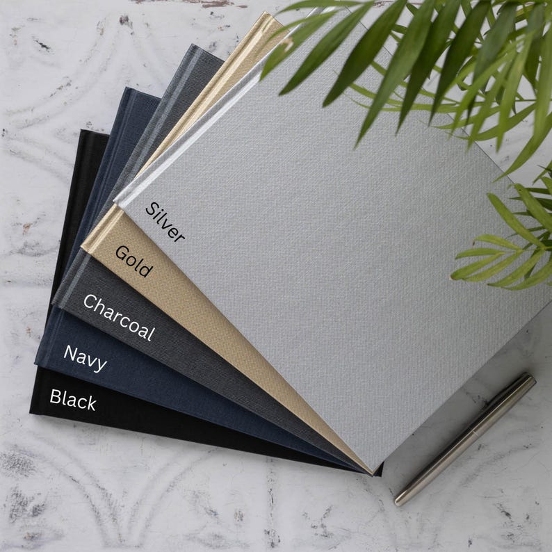 May include: A stack of five notebooks in different colours, including black, navy, charcoal, gold, and silver. The notebooks are arranged in a fan shape, with the silver notebook on top. The notebooks have a textured fabric cover and a simple design.