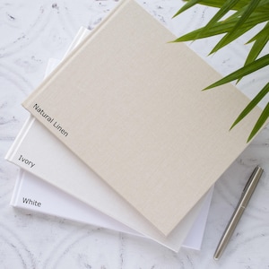 May include: Three guest books in different colours: white, ivory, and natural linen. The books are stacked on top of each other, with the natural linen book on top. The books are on a white marble surface.