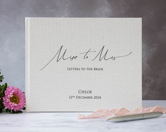 May include: A white linen-covered guest book with the text "Miss to Mrs Letters to the Bride" and the name "CHLOE 12th DECEMBER 2024" printed on the front.