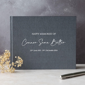 May include: A dark blue linen-textured photo album with the text "HAPPY MEMORIES OF Connor Sean Butler 25th JUNE 1955 - 19th DECEMBER 2024" printed in white.