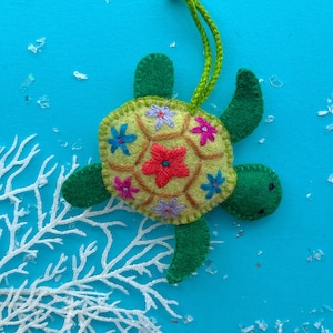 May include: A handmade felt turtle ornament with a light green body and a yellow shell decorated with embroidered flowers in shades of pink, blue, and red. The ornament has a green loop for hanging and is set against a blue background.