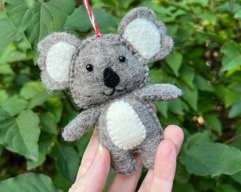 Può includere: Un ornamento di koala in feltro grigio con orecchie in feltro bianco e una toppa di pancia in feltro bianco. L'ornamento è appeso a uno spago rosso e bianco.